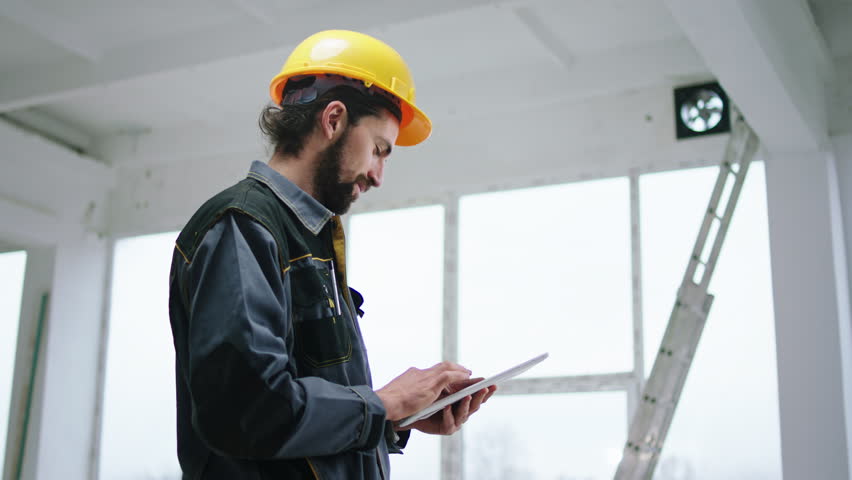Happy constructor worker man holding digital tablet to analysing some work at construction site he wearing special uniform and safety yellow helmet