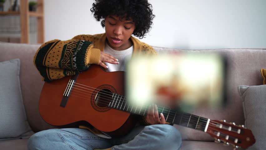 Beautiful american young woman put phone on tripod and records video cover, plays guitar and sings song. Toothy smile, laugh, conversation in video vlog. Teaches or learns to play musical instrument.