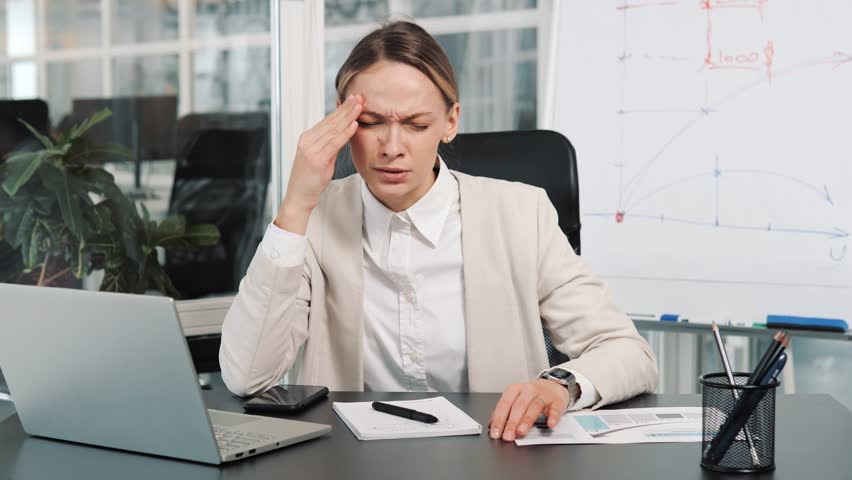 Young exhausted, overworked female office worker sitting in the workplace with a severe headache massaging head. Portrait of unhealthy woman suffering from stress at work, sedentary working computer. Royalty-Free Stock Footage #1100264727