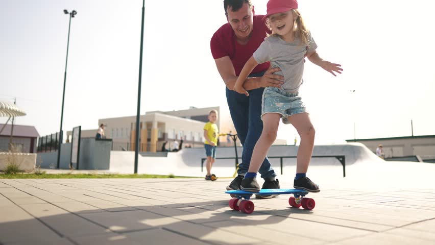 learn to skateboard. dad teaches daughter to ride a skateboard outdoors at the playground. father and daughter play training concept. parent teaching child daughter to skateboard lifestyle Royalty-Free Stock Footage #1100264965