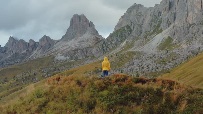Hiker stands exploring nature of high mountain cliffs around Pass Giau. Female tourist stands on top of hill examining vast mountain valley against blue sky