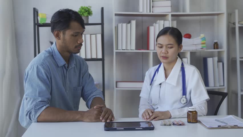 Asian female doctor in white medical coat using clipboard and tablet discussing results or symptoms with male patient sitting at table in health clinic | Shutterstock HD Video #1100266573