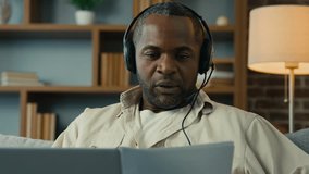 Mature african american man in headphones remote discuss with partner at online conference use laptop at home businessman talk on video call holding documents virtual negotiation concept distance work