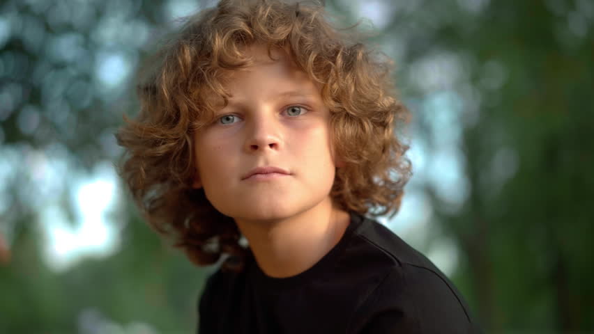 Headshot of curious cute Caucasian boy looking around sitting outdoors at background of green spring summer tree crowns. Close-up portrait of charming child enjoying leisure outdoors and posing