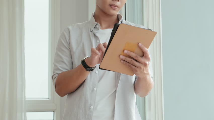 Tilt up portrait shot of young Asian man standing at home, using digital tablet and then posing for camera