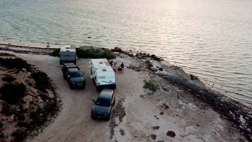 Aerial view pulling away from RV trailers parked along Australia's coastline at sunset.