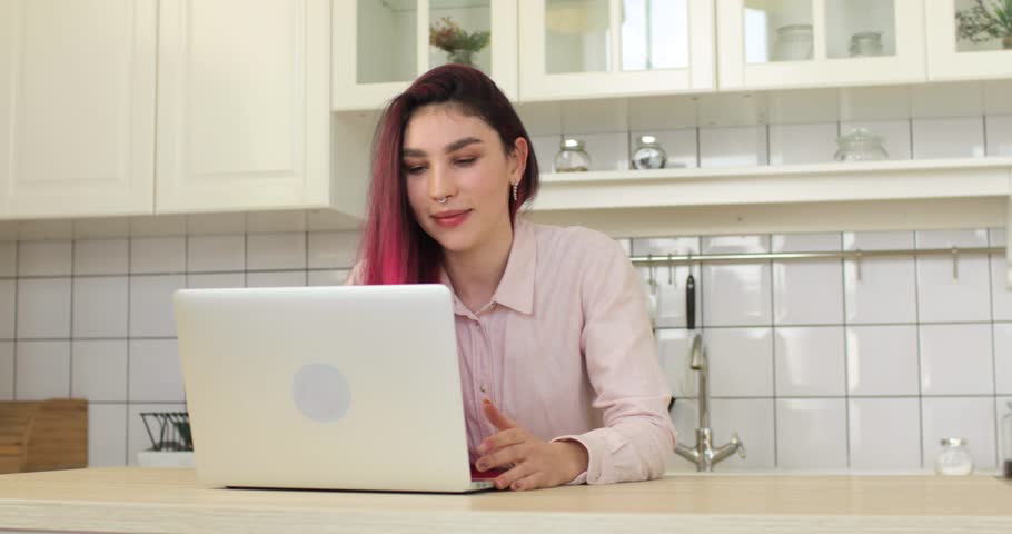 Portrait of young woman with pink hair, using laptop computer in the kitchen. She is watching news on laptop screen and working or studing at home workplace. Home office concept.