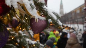 Heart shaped Christmas bauble hangs on spruce tree branch covered with snow. Soft focus. Blurred people walking by Moscow Kremlin in the background. Handheld real time video. Winter holidays in Russia