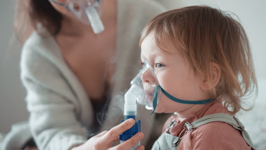 Asian mother helping sick daughter use nebulizer while embracing her on couch at home. Woman makes inhalation with equipment to toddler boy. Ill child lying on couch having respiratory illness helped | Shutterstock HD Video #1100293467
