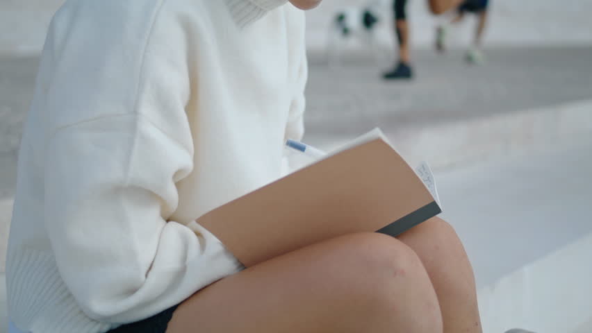 Unknown young woman writing into notebook sitting staircase close up. Girl hands with pen making notes in notepad diary outdoors. Unrecognizable freelancer working sitting on stairs. Urban lifestyle.