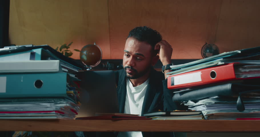 Busy stressed black male finance manager, CEO executive works hard at office table full of paperwork checking with notes | Shutterstock HD Video #1100299595