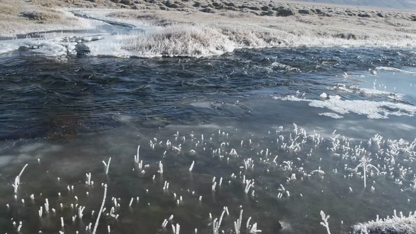 Fast-moving stream of narrow river runs along frozen banks on sunny day. Landscape of autumn nature with melting ice islands and frosted grass in highland