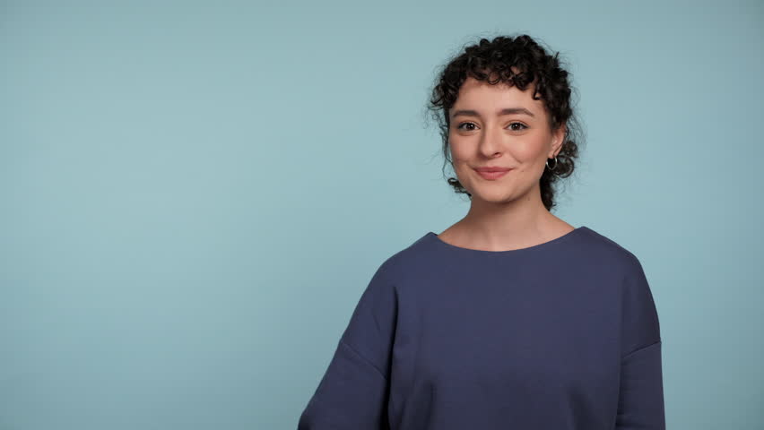 Smiling young curly woman holds bitcoin in hand, looking at camera shows like with hand thumbs up, symbol of cryptocurrency. Portrait positive female standing on isolated light blue background