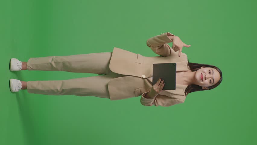 Full Body Of Asian Business Woman Smiling While Using And Pointing To Tablet On Green Screen Background In The Studio
