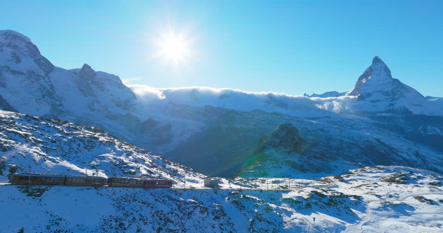 Aerial drone footage of Gornergrat bahn railway running towards summit station with Matterhorn mountain peak background in Zermatt on a sunny winter day. Swiss Alps, Switzerland travel journey trip.