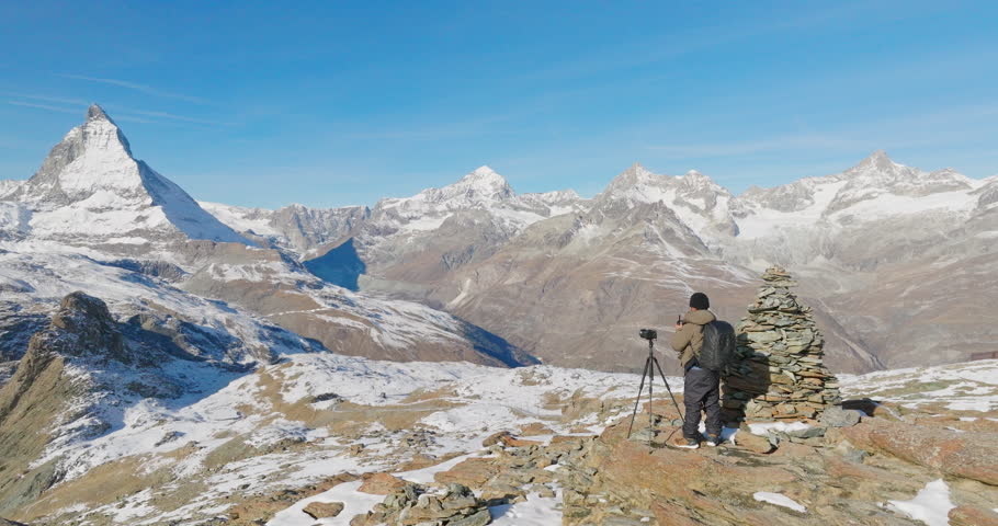 Young man backpacker take a photo by camera at Switzerland mountain peak view point with iconic famous landscape Matterhorn background. Aerial drone orbit shot, Nature, Travel and Adventure concept.