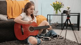 Young teenager girl with guitar making live stream to followers and explaining online how to play musical instrument. Pretty female guitaris making video on camera for social media channel
