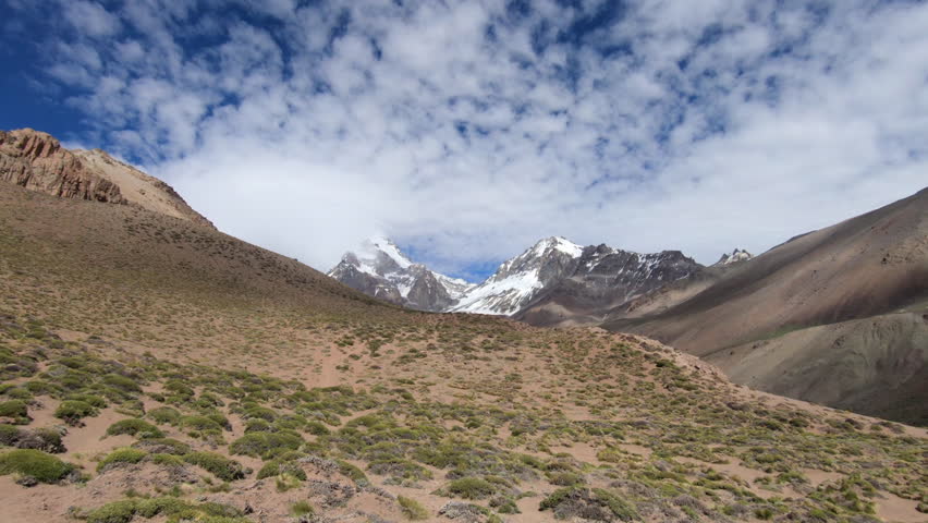 View of the summit of Aconcagua from the approach to basecamp, with blue sky and clouds moving over the mountains creating shadows in the valley