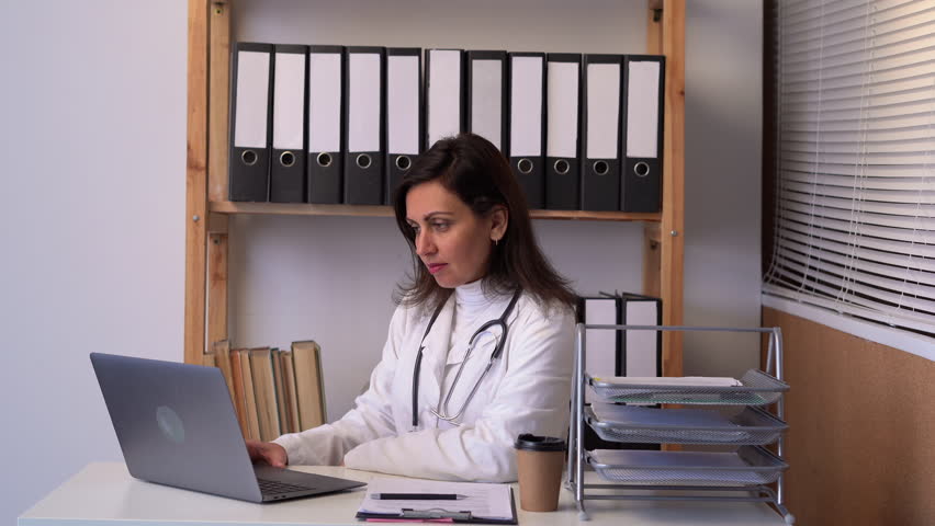 Telemedicine concept. Indian female doctor talking with patient using laptop online video webinar consultation while working in clinic office. | Shutterstock HD Video #1100313147