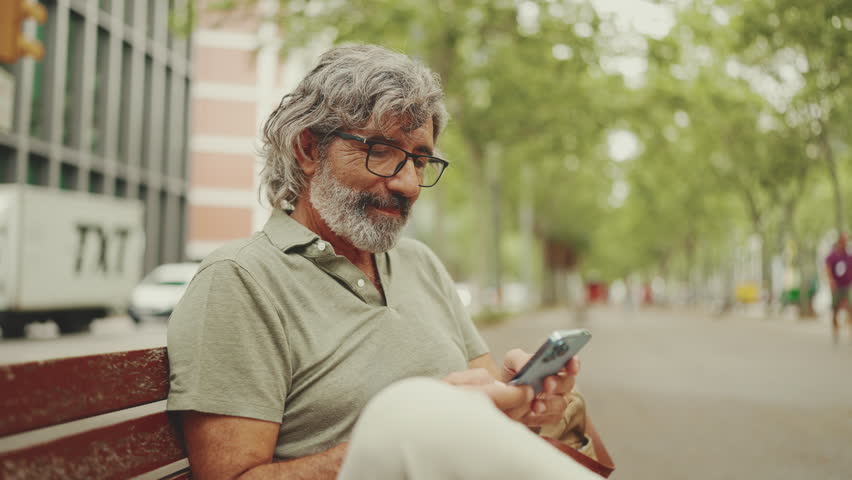 Positive middle-aged man with gray hair and beard wearing casual clothes sits on bench and uses mobile phone. Mature gentleman in eyeglasses raises his head, looks at the camera and smiles