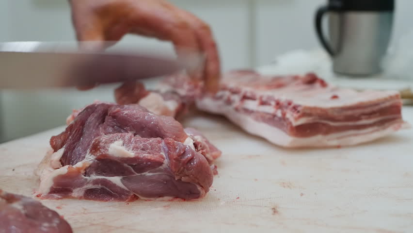 The process of cutting fresh raw meat by a butcher on a cutting board in a butcher shop using a large sharp knife. Hands of a male butcher holding a knife cut raw meat into pieces. Retail meat trade Royalty-Free Stock Footage #1100316025