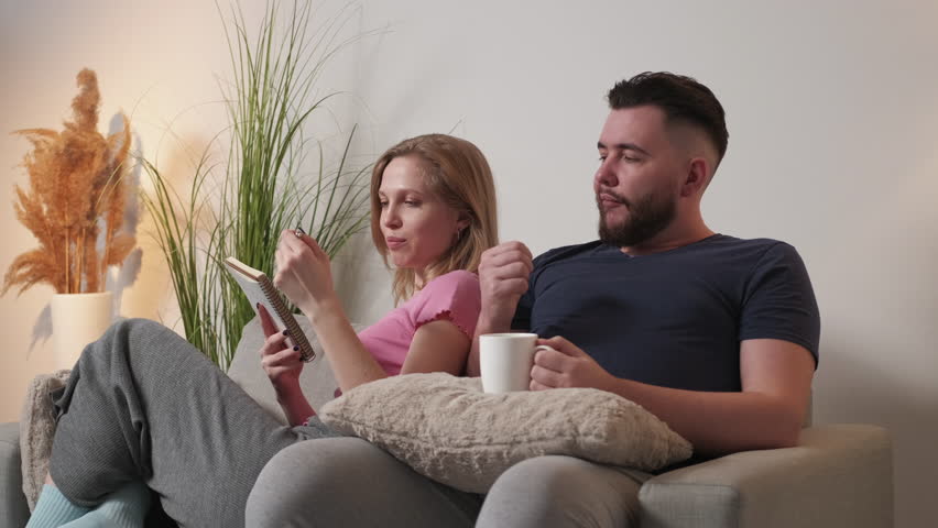 Planning list. Family couple. Home leisure. Funny man counting fingers while woman making notes in copybook sitting sofa together in light room interior. Royalty-Free Stock Footage #1100316403