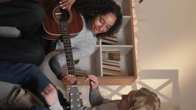 Vertical video. Guitar playing. Home lesson. Beloved couple. Inspired man showing woman accords on acoustic instrument sitting light room interior.