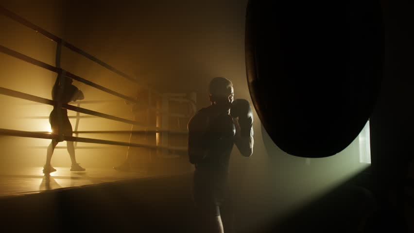 Friends training and kicking punching bag in dark, modern fitness club. Man and woman working out with a coach to gain muscle strength and endurance. High quality 4k footage Royalty-Free Stock Footage #1100319109
