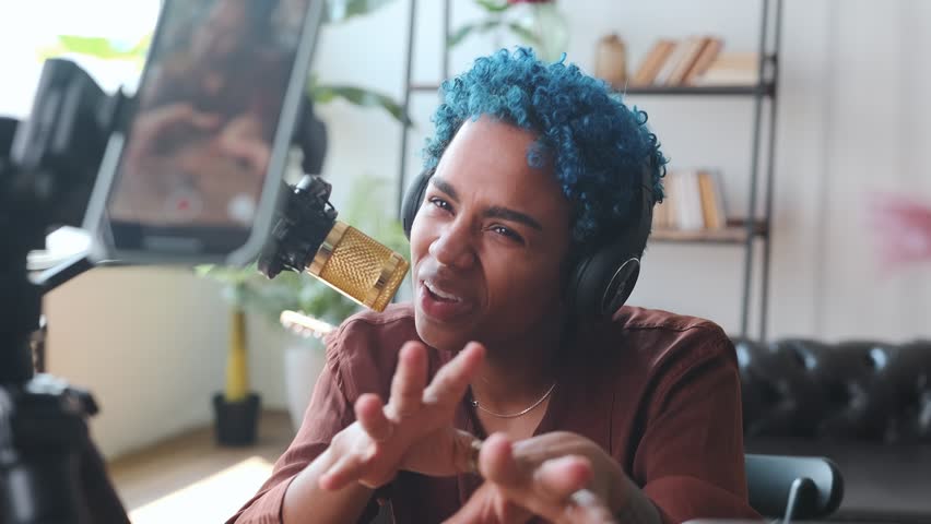 Young confident eccentric African American woman blogger recording video podcast on mobile phone camera mounted on tripod making career as influencer and pushing back microphone sits by table Royalty-Free Stock Footage #1100324235