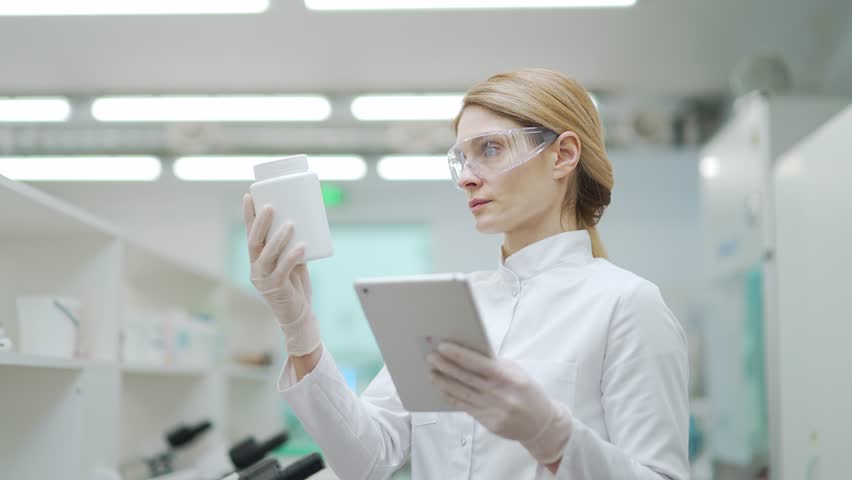 Focused mature woman scientist researcher with protective glasses looking at drug medicine product and compares the research in digital tablet at microbiology pharmaceutical biochemistry laboratory