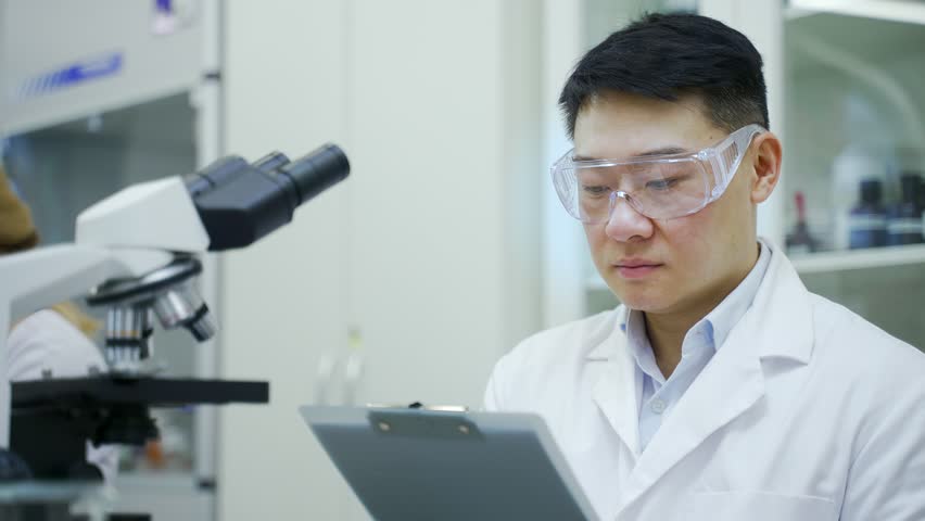 Concentrated mature asian man scientist with protective glasses and gloves working make notes descriptions after research with microscope at microbiology pharmaceutical biochemistry laboratory indoors Royalty-Free Stock Footage #1100329141