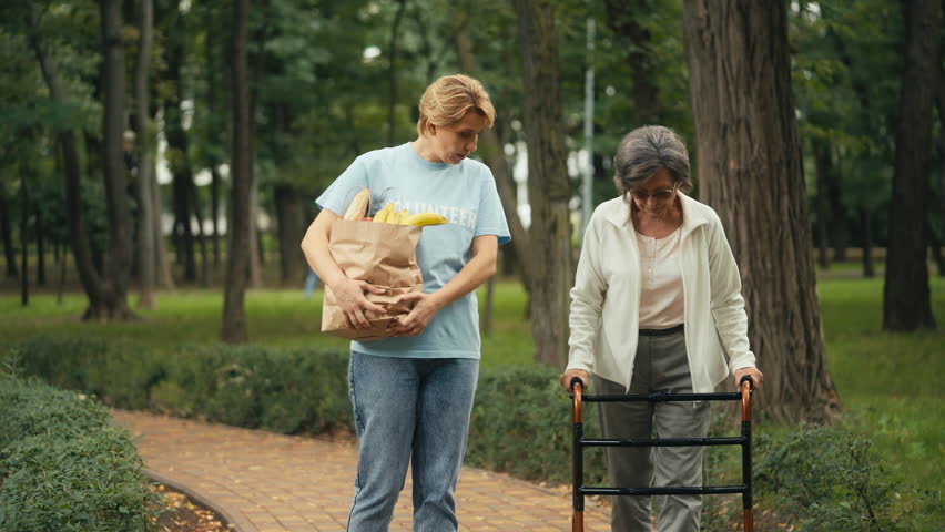 Female volunteer carrying grocery bag, supporting elderly woman with disability Royalty-Free Stock Footage #1100334567