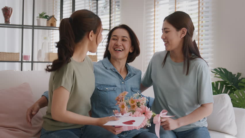 Mother's day two grown up child cuddle hug give flower gift box red heart card to mature mum. Love kiss care mom asia middle age adult three people sitting at home sofa happy smile enjoy family time. Royalty-Free Stock Footage #1100336653