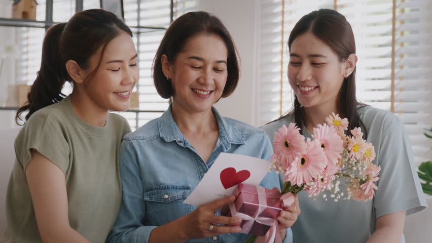 Mother's day two grown up child cuddle hug give flower gift box red heart card to mature mum. Love kiss care mom asia middle age adult three people sitting at home sofa happy smile enjoy family time. Royalty-Free Stock Footage #1100336655