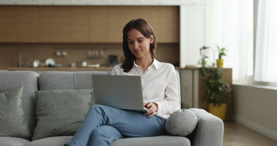 Happy young freelance business woman working at home, typing on laptop, using online app, sitting on sofa, holding computer on lap, smiling, laughing. Internet communication Royalty-Free Stock Footage #1100336951