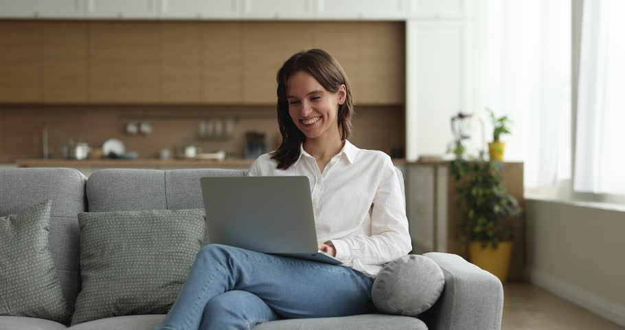 Happy young freelance business woman working at home, typing on laptop, using online app, sitting on sofa, holding computer on lap, smiling, laughing. Internet communication | Shutterstock HD Video #1100336951