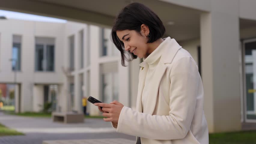 Young businesswoman working from her phone sitting outside an office building in a financial center. Millennial person using a smartphone outdoors. Girl chatting with her colleagues on her cell. | Shutterstock HD Video #1100338975