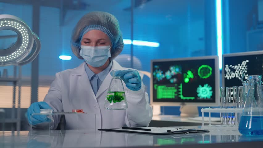 A female scientist adds brown powder to a clear liquid in a flask. The woman observes the reaction of the sample, stirs the liquid and writes the test data. Research in a modern biochemical laboratory | Shutterstock HD Video #1100339539