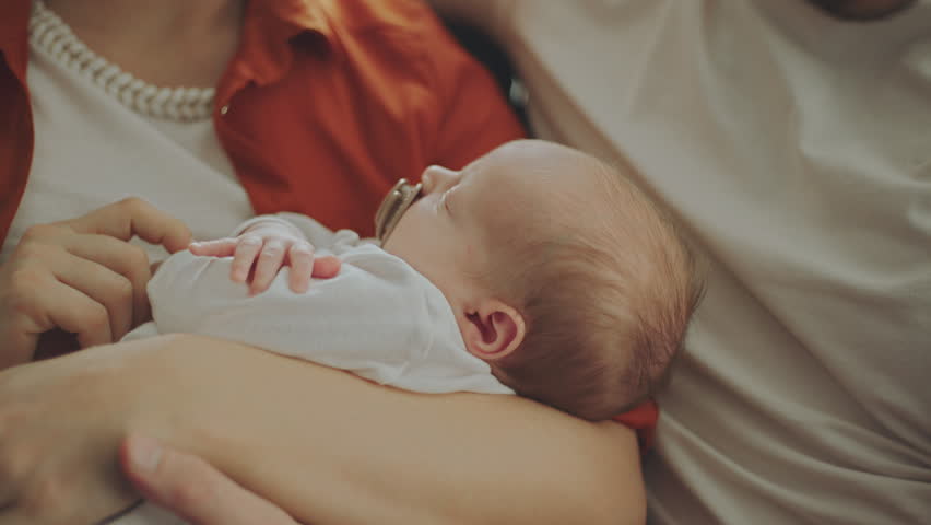 Close-up shot of an adorable sleeping newborn baby with parents sitting on the sofa at home in living room. Mom taking off the pacifier from baby mouth in slow-motion | Shutterstock HD Video #1100340647