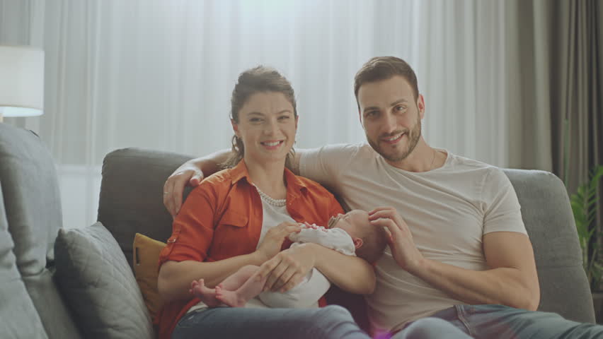 Beautiful young family with a newborn baby in their arms looking at camera and smiling while sitting on the sofa Royalty-Free Stock Footage #1100340653