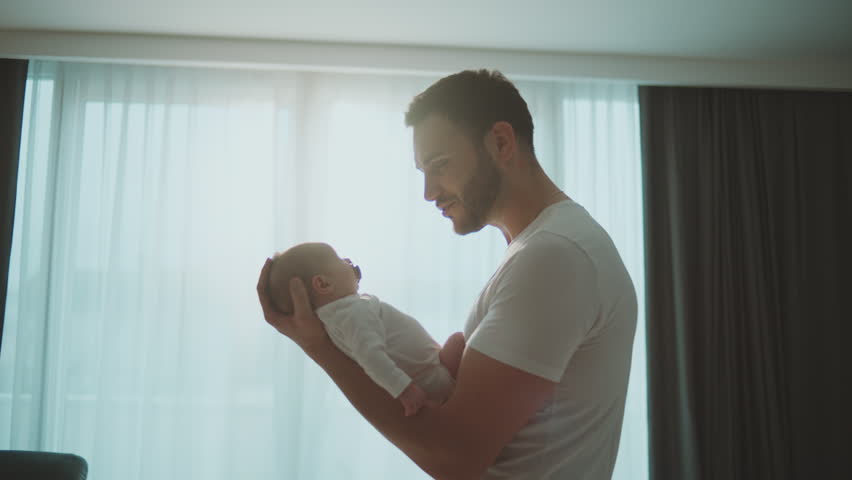 Medium shot of a young father holding his newborn baby in arms, staring at baby and talking with him against large window. Slow-motion Royalty-Free Stock Footage #1100340675