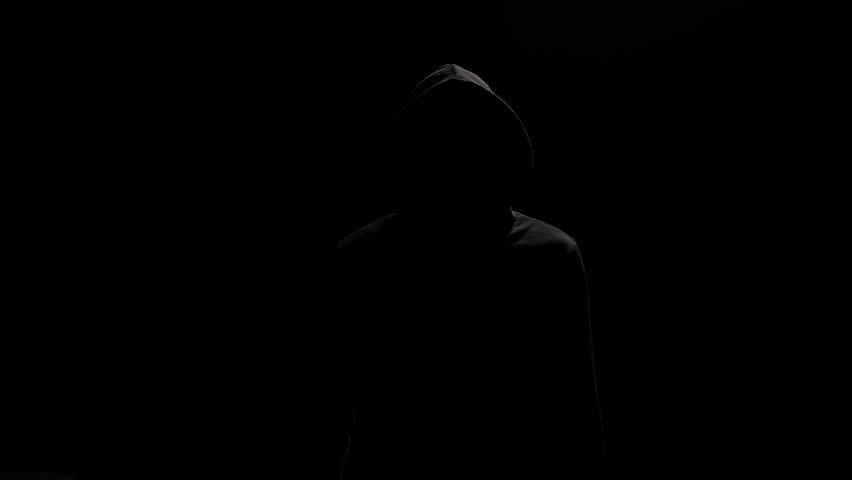 Isolated portrait mysterious figure in black hoodie, pointing at camera finger. Figure of man in black hoodie emerges from the shadows, directing pointed finger at lens. Black-clad stranger in backlit Royalty-Free Stock Footage #1100342799