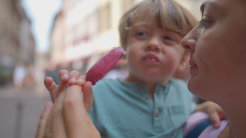 Mother sharing ice cream with son during summer day. Woman eating sweet dessert snack holding child in street while snacking