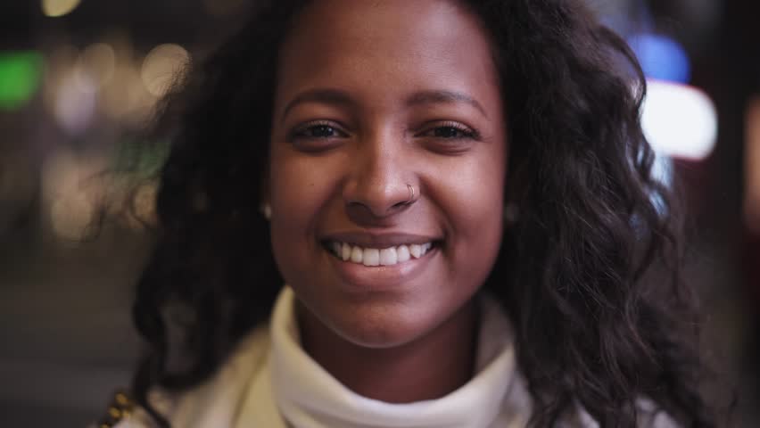 Pretty young African woman smiling looking at camera in the urban street. Gorgeous black girl is shown happy in a close-up shot at night. Cheerful people sightseeing in the city.  | Shutterstock HD Video #1100349157
