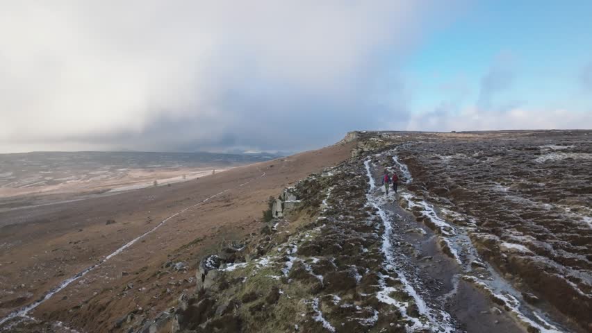 Two people hiking along a frozen moorland path on top of a rock formation in the Peak District.