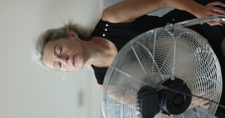 Middle-aged woman exhausted by unbearable heat, cooling herself using electric floor fan sit on sofa in living room. Extreme hot weather, need air-condition, climate control system, hormonal imbalance Royalty-Free Stock Footage #1100353633