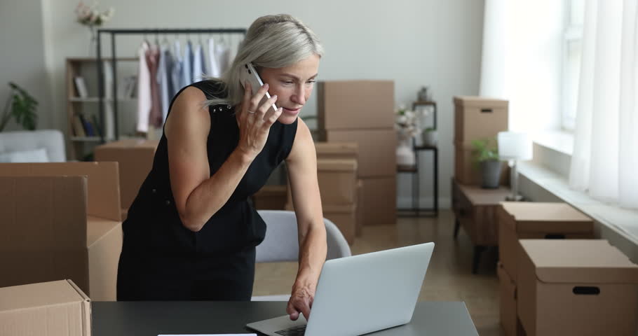 Mature pretty woman use laptop working and talking on smart phone, make transportation company services order at relocation day, speaks to client sell goods working in own office with cardboard boxes Royalty-Free Stock Footage #1100353637