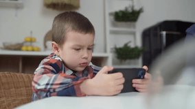 Cute Little Boy Sitting At Table With Phone In His Hands, Plays Mobile Games, Watches Videos and Social Media