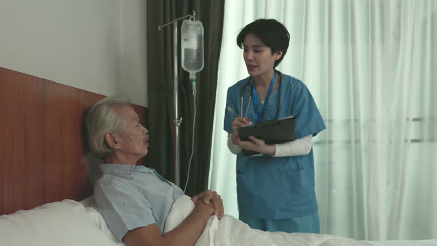 An elderly patient is lying in a hospital bed while a young doctor or nurse dressed asks about their symptoms. The doctornurse listens attentively and provides medical advice to the patient. | Shutterstock HD Video #1100357037