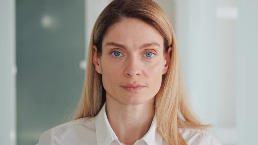 Head shot close up portrait of serious focused millennial dreamy successful business woman looking at camera. Female leader have natural makeup, blue eyes dreseed in white shirt standing in office. | Shutterstock HD Video #1100357753